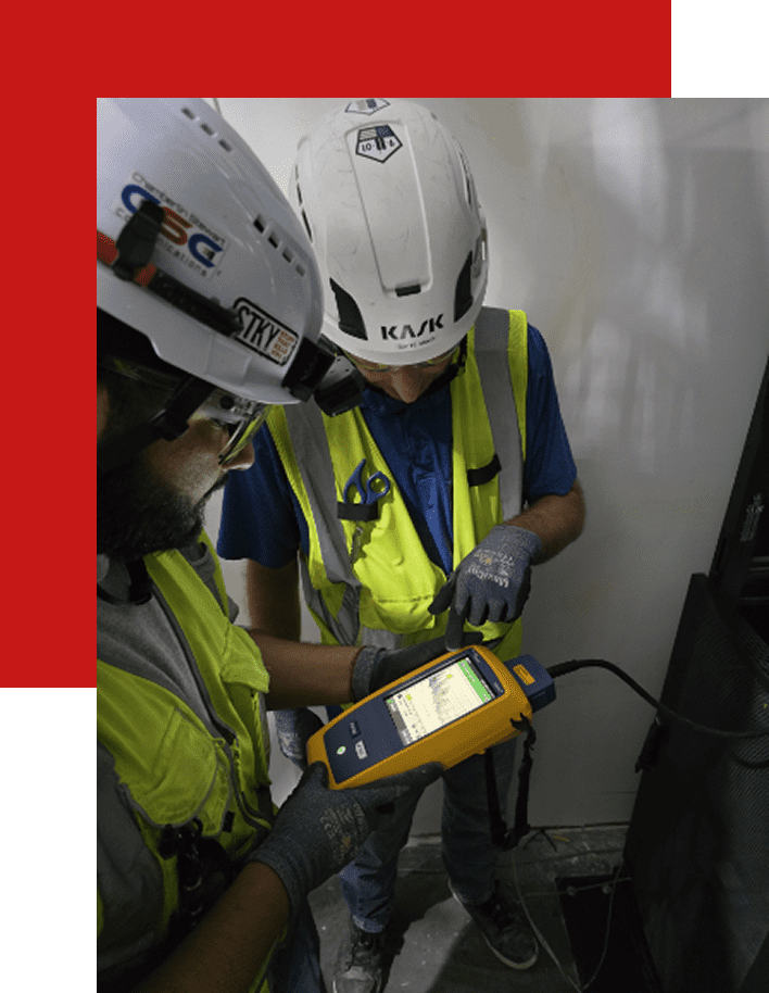 Two workers in safety gear using a handheld device indoors.